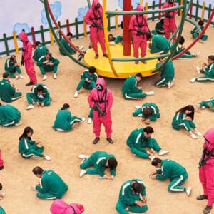 Squid Game: Players in green suits sit on sand, watched by guards in pink suits with masks, some on a playground structure.