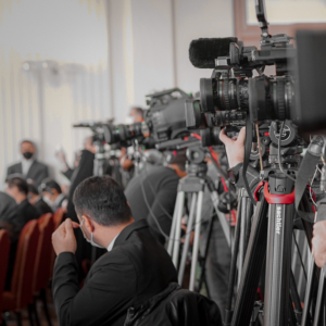 Media frenzy: Cameras focus on politicians' private lives. Press conference with masked attendees, suggesting antisocial media scrutiny.