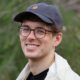 Daniel Finlay portrait. Man with glasses, baseball cap, and white jacket smiling outdoors.