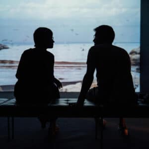 Silhouetted people listening, expanding worldview. Two figures converse on a bench, ocean backdrop. Listening to people we disagree with.