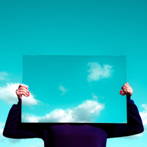 Sky reflected in mirror held by person, exploring identity. Blue sky, clouds, and purple sweater create a surreal, ethical, visual metaphor.