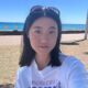 Ariel Bai selfie. Young woman with sunglasses on her head, standing near a beach with blue water and clear sky. She's wearing a white t-shirt.