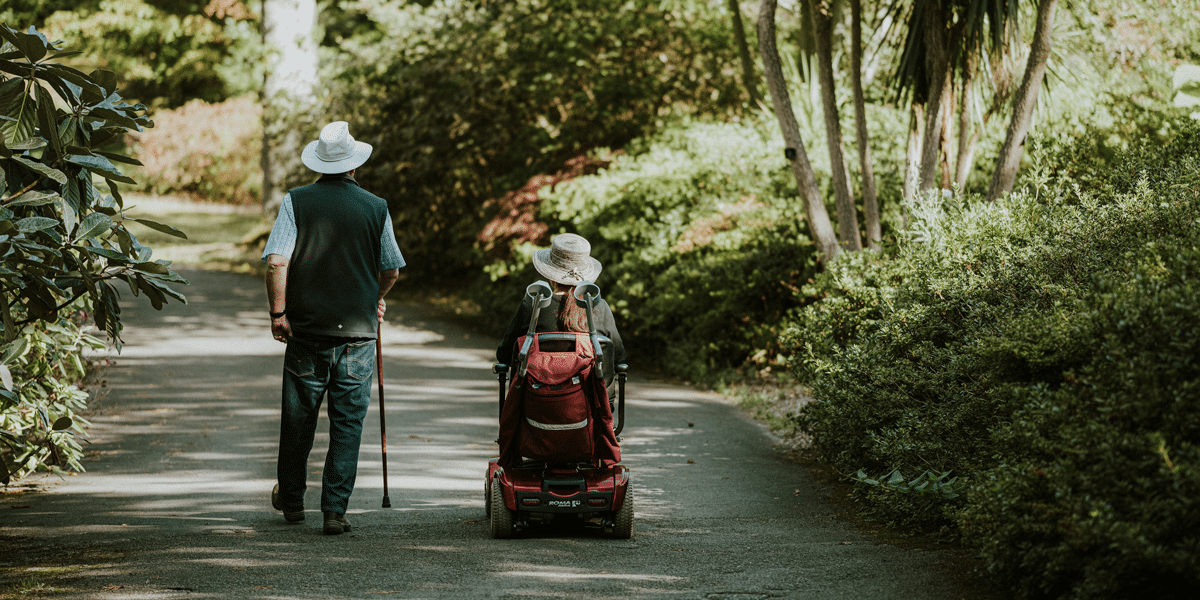 Duties of care: Person with mobility scooter and companion walking on a path in a lush garden. Care and support in older age.