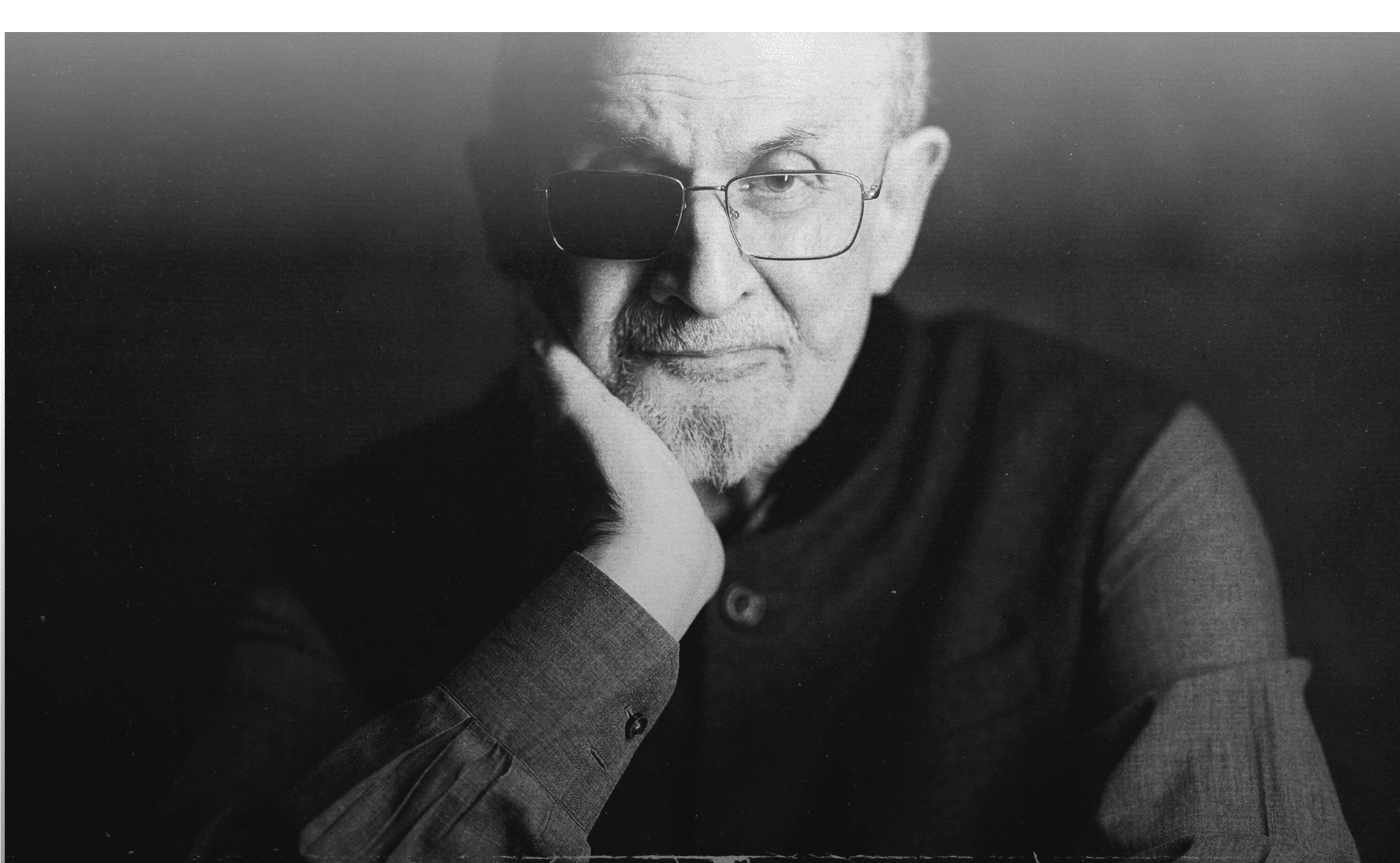 Black and white portrait of Salman Rushdie, wearing glasses with one lens blacked out, resting his chin on his hand, looking at the camera.
