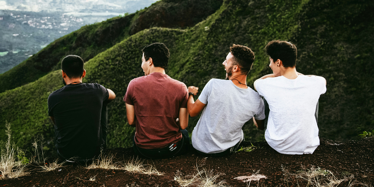 Four friends, seen from behind, sit atop a hill overlooking a lush valley, embodying the 'good guy' isn't always the kind person concept.