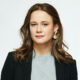 Auto Draft: Portrait of a woman with wavy brown hair, wearing a gray shirt, black blazer, and large gold earrings against a white background.