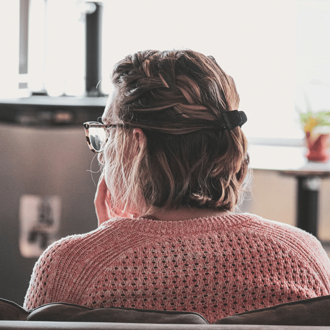 Back view of a thoughtful teacher with glasses, hair in a braid, and pink sweater. Moral injury and compassion concept.