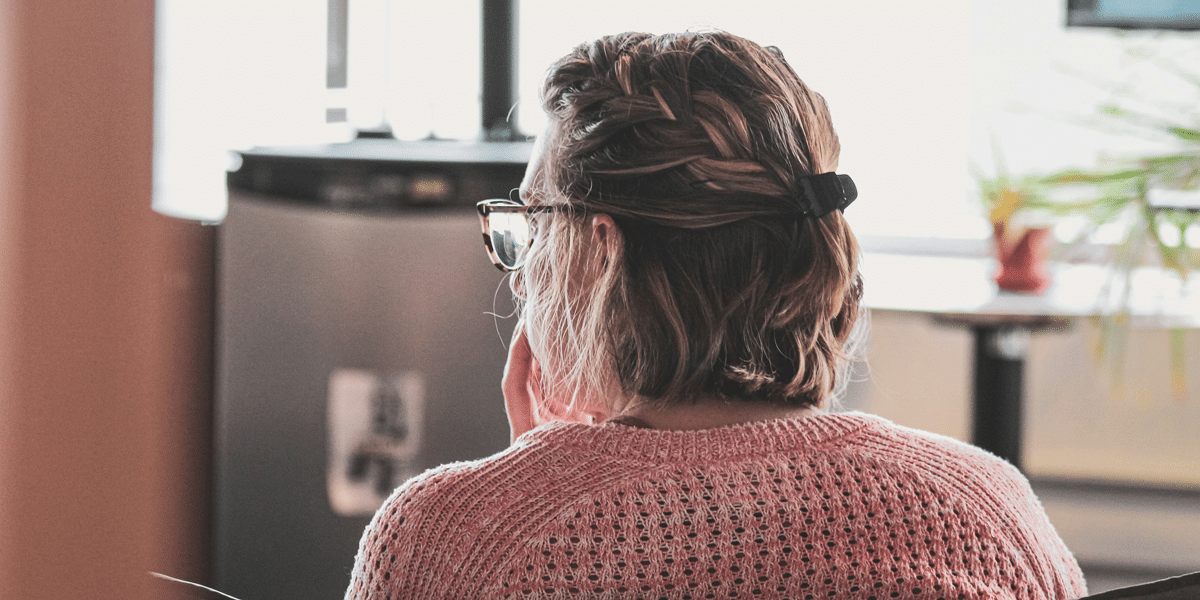 Teacher with moral injury, seen from behind, wearing glasses and a pink sweater, hair braided, contemplating compassion.