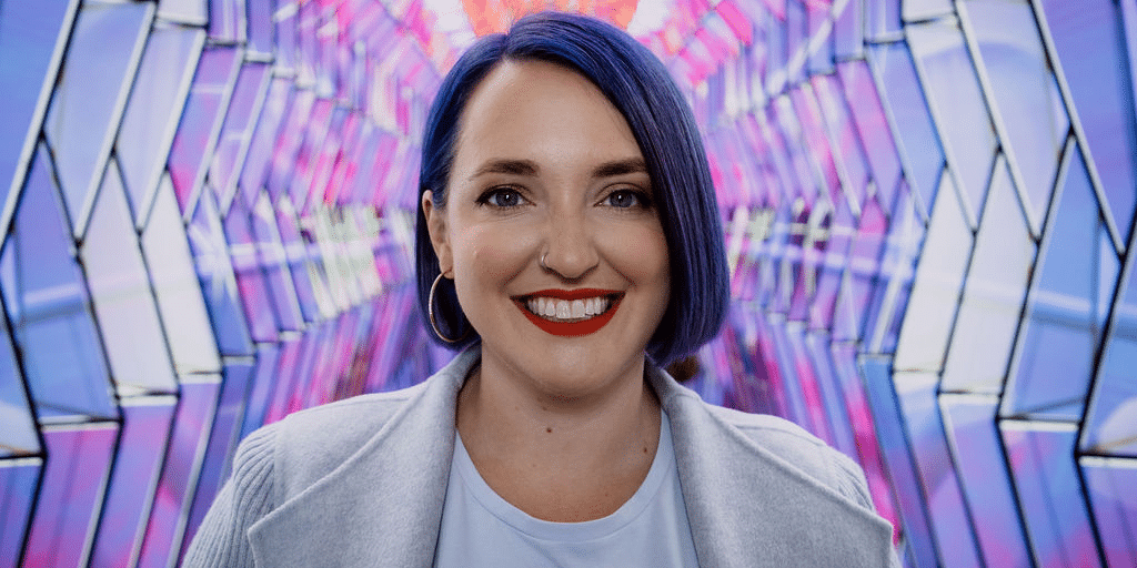 Aubrey Blanche, director of responsible leadership, smiles against a colorful geometric background. She has blue hair and red lipstick.