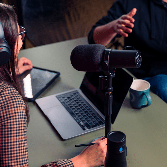 People recording a podcast, discussing media literacy and platforming. Microphone, laptop, tablet, and water bottle on a green table.