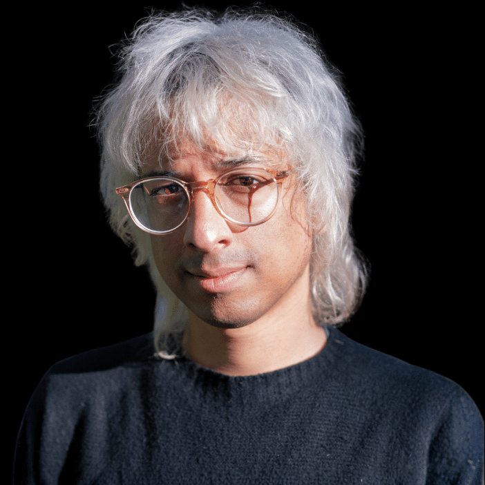 Aaron Cornelius portrait. Man with glasses, black sweater, and white hair on a black background.