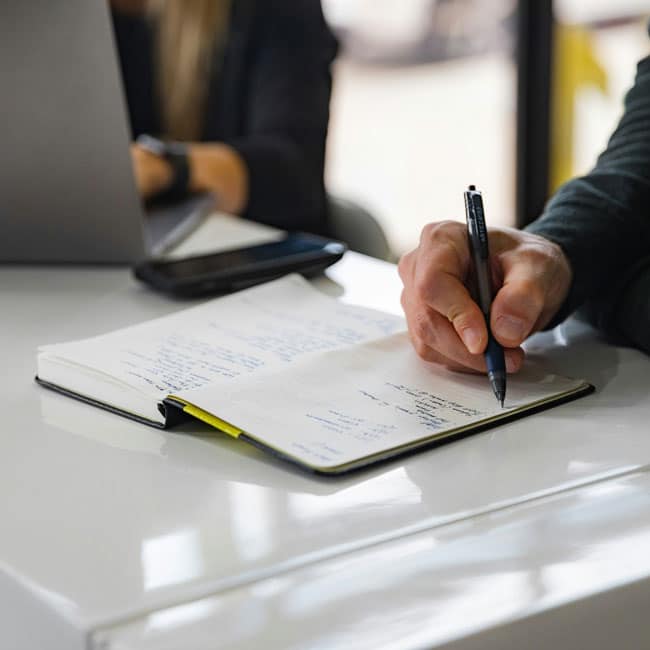 Taking notes. Hand writing in notebook with pen, laptop and phone visible. Is an AI notetaker in the room?
