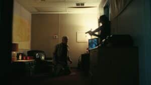 Tense political stand-off: A man kneels, facing a figure holding a gun in a dimly lit room, illustrating resistance in one battle after another.