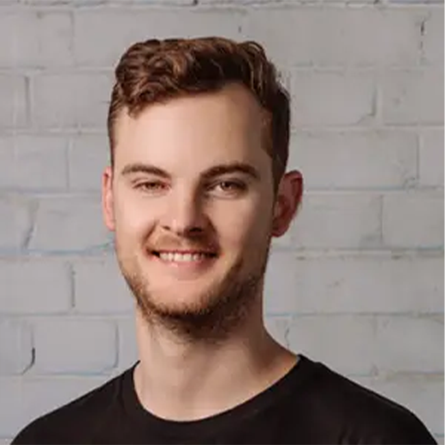 The Ethics of Bans: Smiling man with short brown hair and a black t-shirt, posing in front of a white brick wall, looking at the camera.