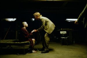 A scene depicting conspiracy theories: a bald woman sits, connected to wires, while a man in a suit holds her hand in a dimly lit room.