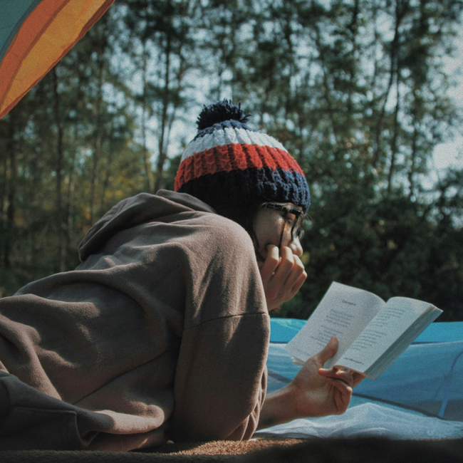 A person wearing a beanie and glasses reads a book inside a tent, surrounded by trees. Reflecting on cultural diversity in literature.