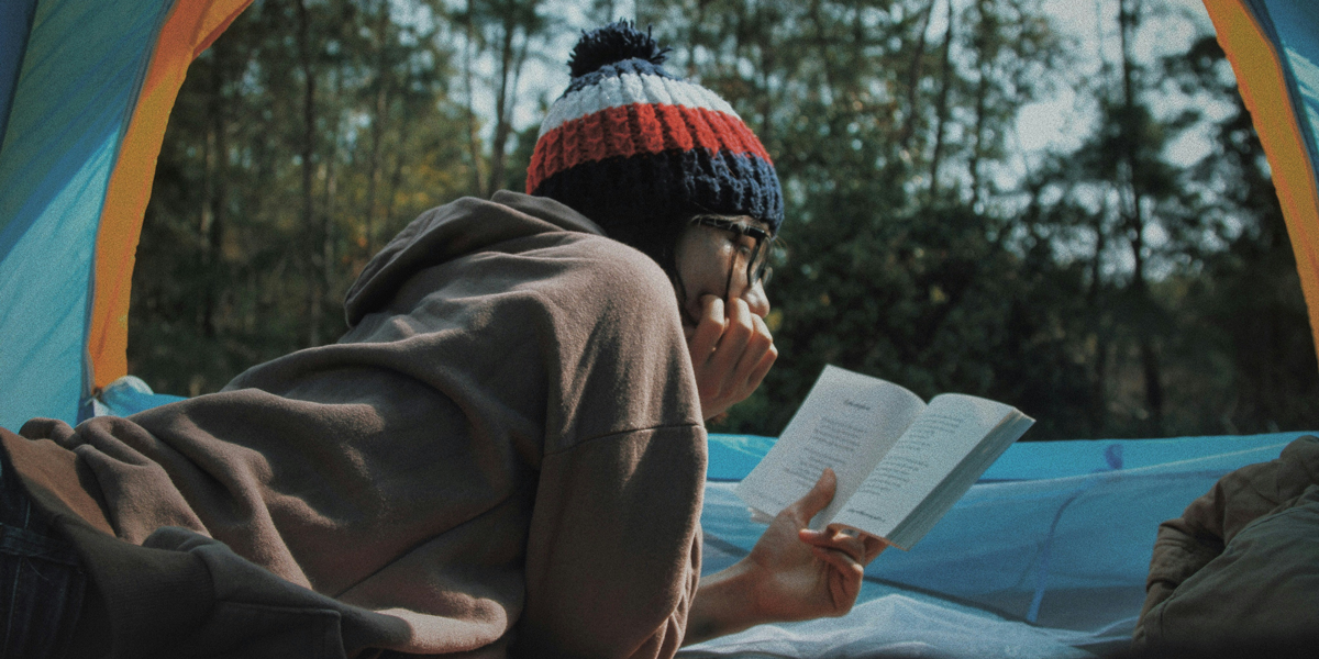 Inside a tent, an individual wearing a beanie thoughtfully reads a book, possibly exploring themes of cultural diversity in literature.