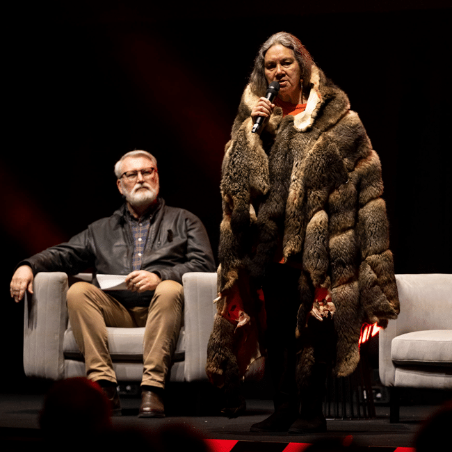 Welcome to Country: Indigenous speaker in possum skin cloak addresses audience, respecting culture. Man sits nearby. Deep respect for traditions.
