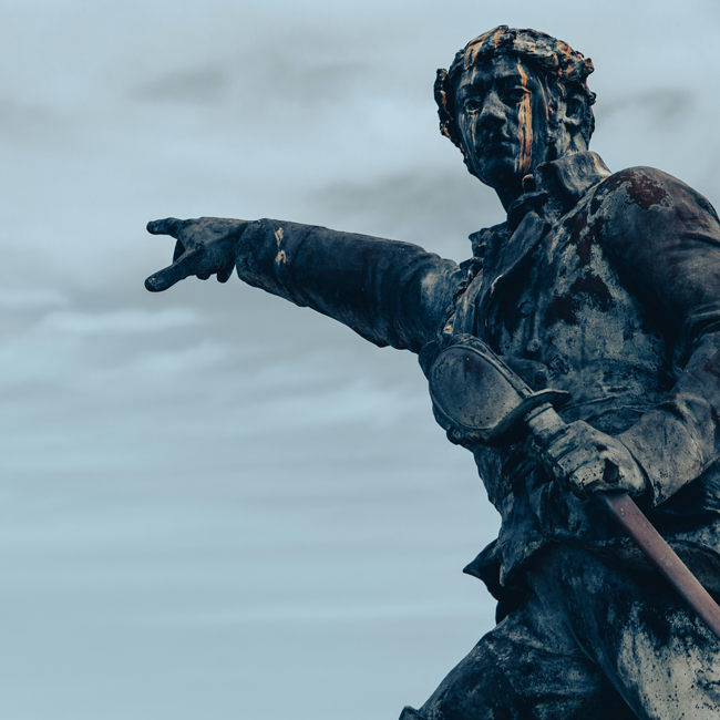 Weathered bronze statue of a man in historical military attire pointing forward with a sword against a cloudy grey sky view