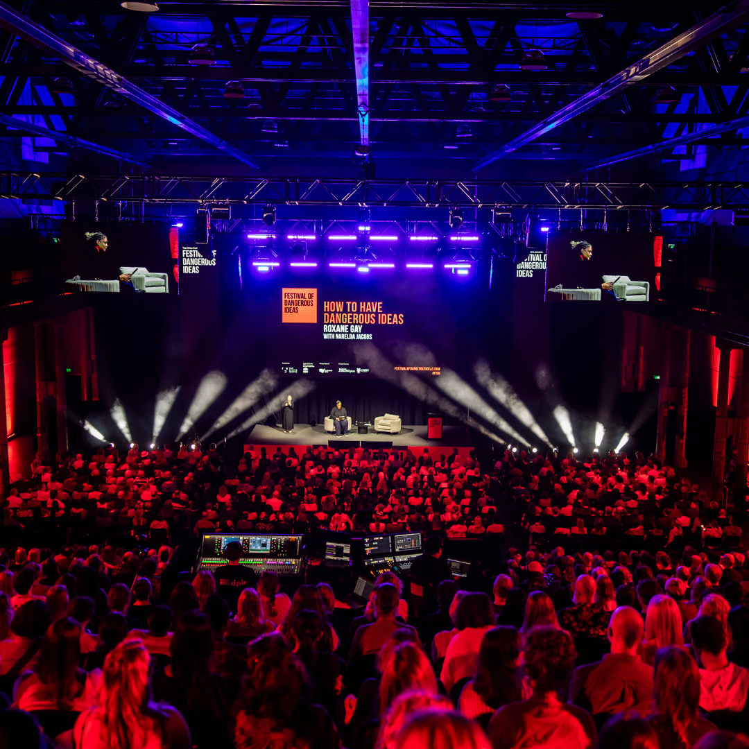 Wide view of a large, dark theater with a brightly lit stage featuring a speaker at the Festival of Dangerous Ideas event