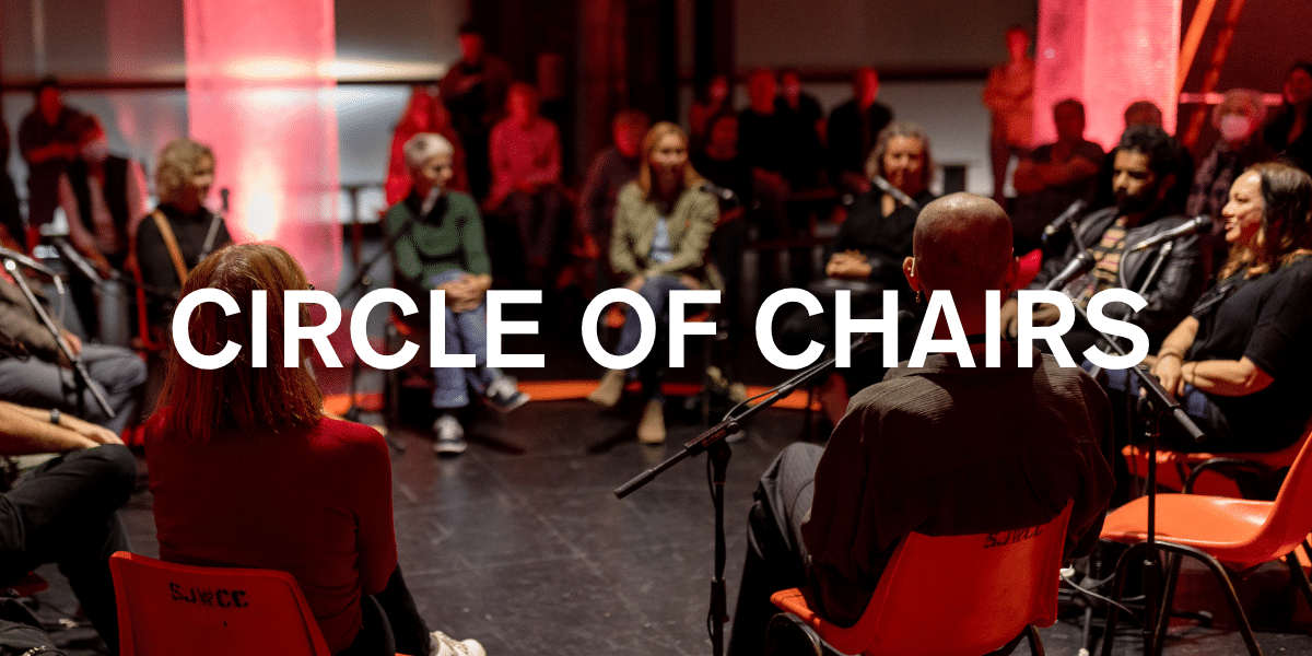 People sit in a circle of chairs during a workshop or performance with microphones and warm red lighting in a dark theater