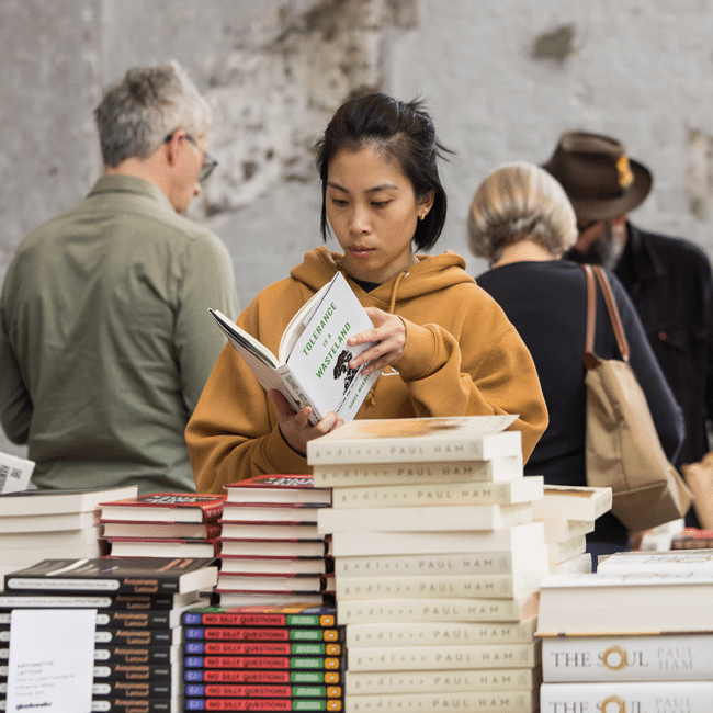 Focused young person in tan hoodie reads Tolerance is a Wasteland book at a market filled with stacks of dangerous reads