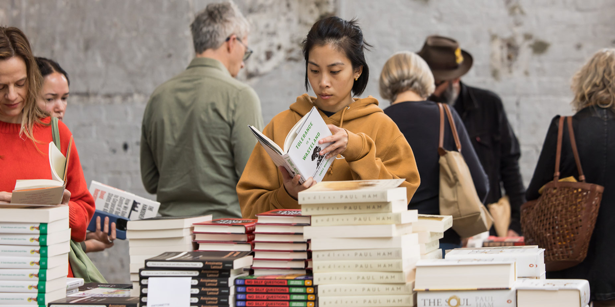 Visitor in a mustard sweater inspects a book at a bustling market display of dangerous reads for Festival of Ideas patrons