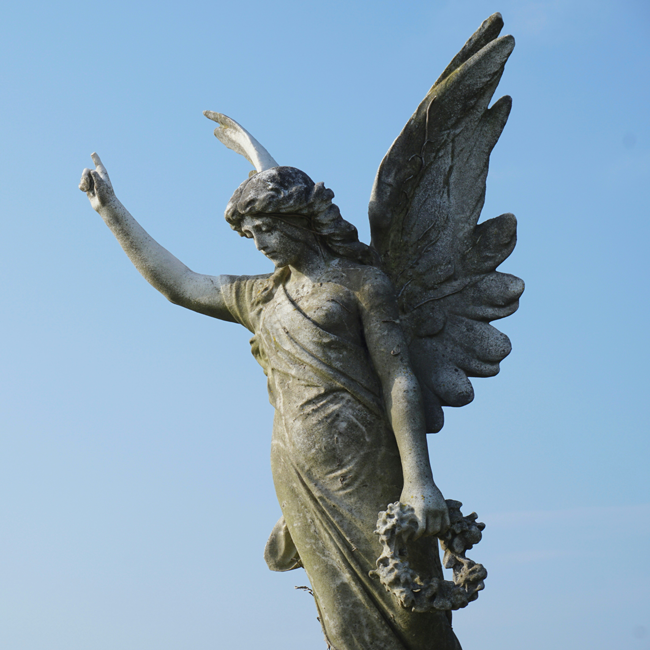 Weathered stone statue of an angel with large wings pointing upward while holding a floral wreath against a bright blue sky