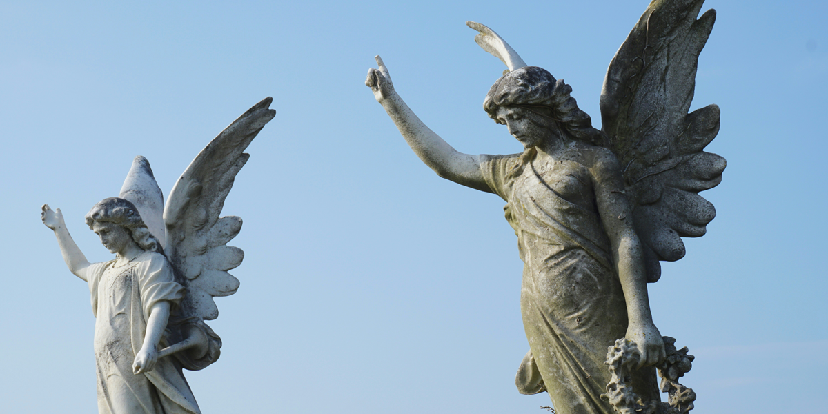 Two aged marble angel grave markers stand against a clear sky representing moral certainty and the heavy burden of ruin