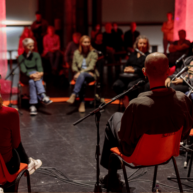 A group of people sit in a circle on stage during a panel discussion, with microphones and an audience watching in the dark.