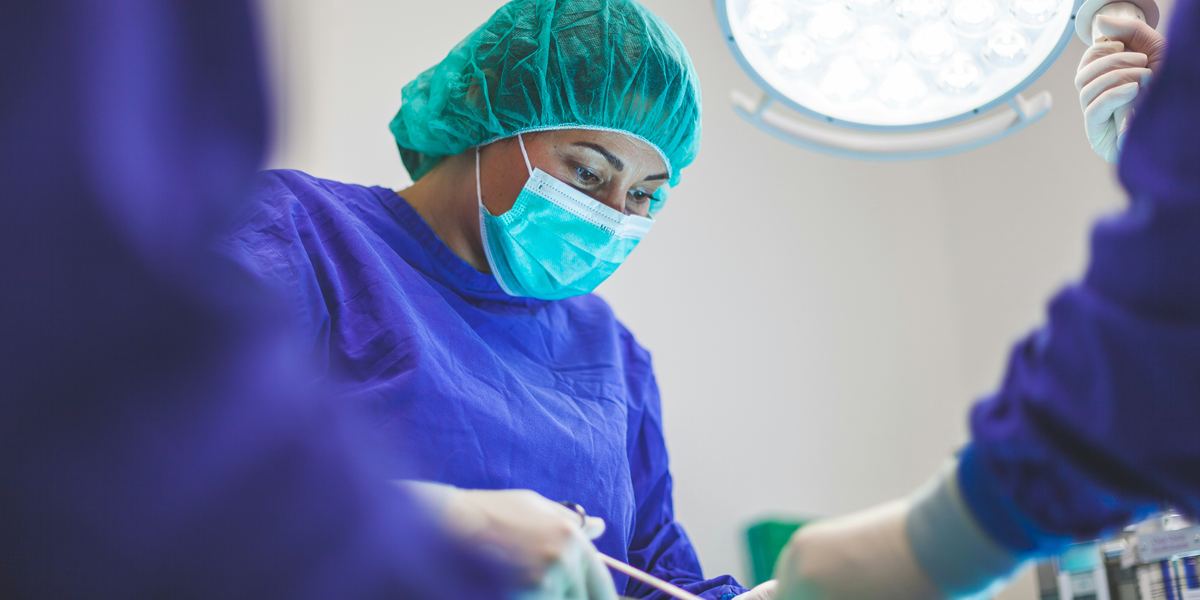 Close-up of a focused female surgeon in blue scrubs and green cap performing an operation under a surgical light.