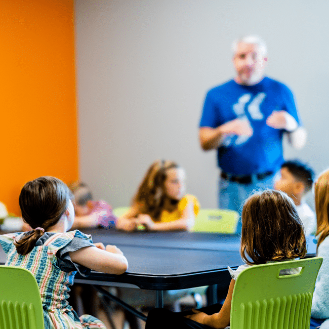 A teacher stands before a group of young children sitting at a table during an ethics education lesson in a bright classroom