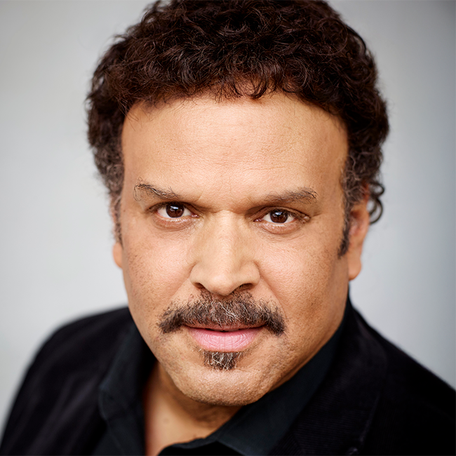 Close-up studio portrait of a serious man with curly hair and a goatee, relevant to the ethics of intervention topic