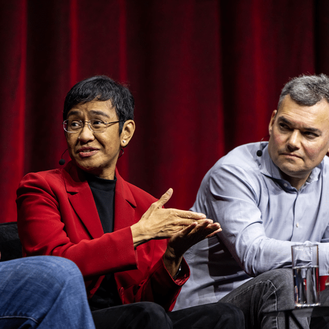 A woman in a red blazer speaks at the Festival of Dangerous Ideas 2026, gesturing while a man sits listening beside her.