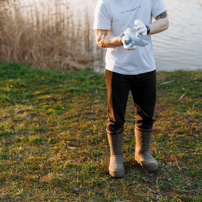 Person in white t-shirt, dark pants, and rubber boots standing outdoors on grass, holding a plastic bag while doing the wrong thing