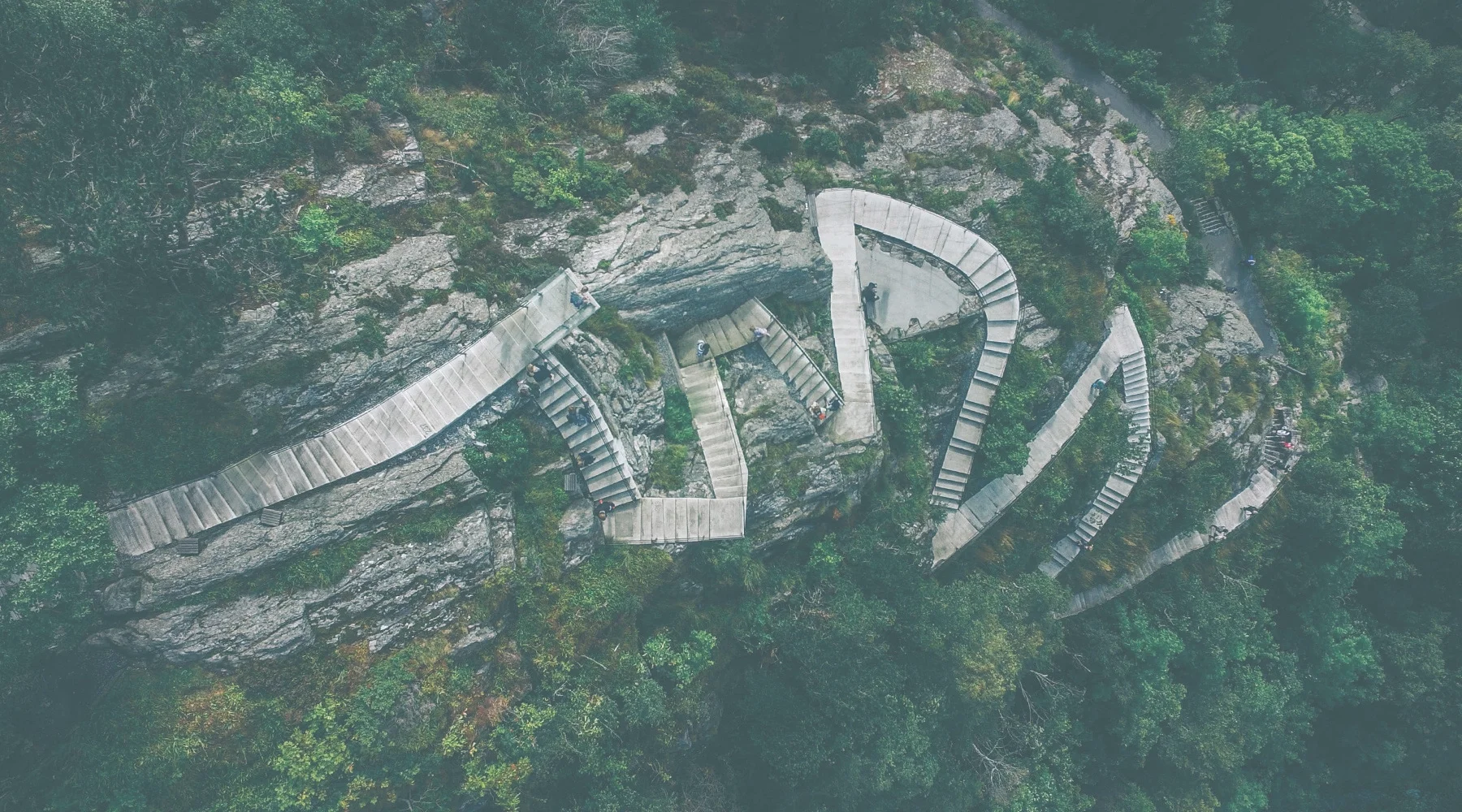Media: Aerial view of winding stairs climbing a rocky cliff face surrounded by lush green trees, showcasing a unique architectural design.