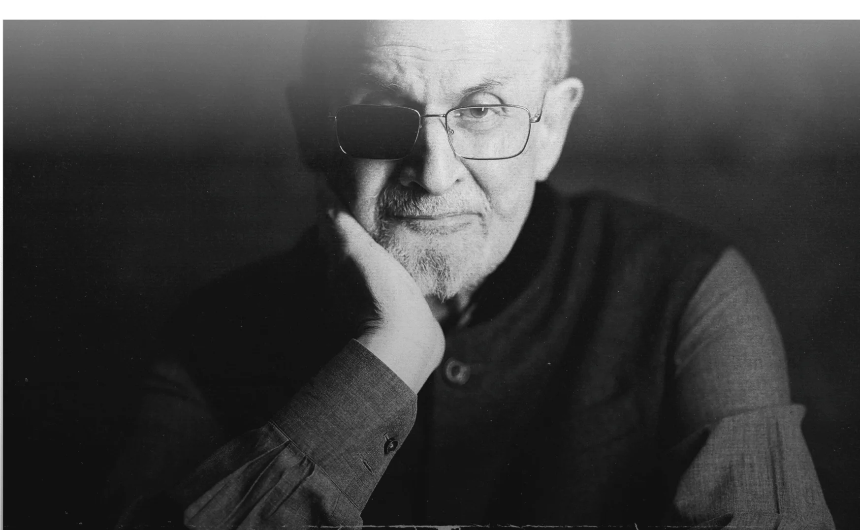 Black and white portrait of Salman Rushdie, wearing glasses with one lens blacked out, resting his chin on his hand, looking at the camera.