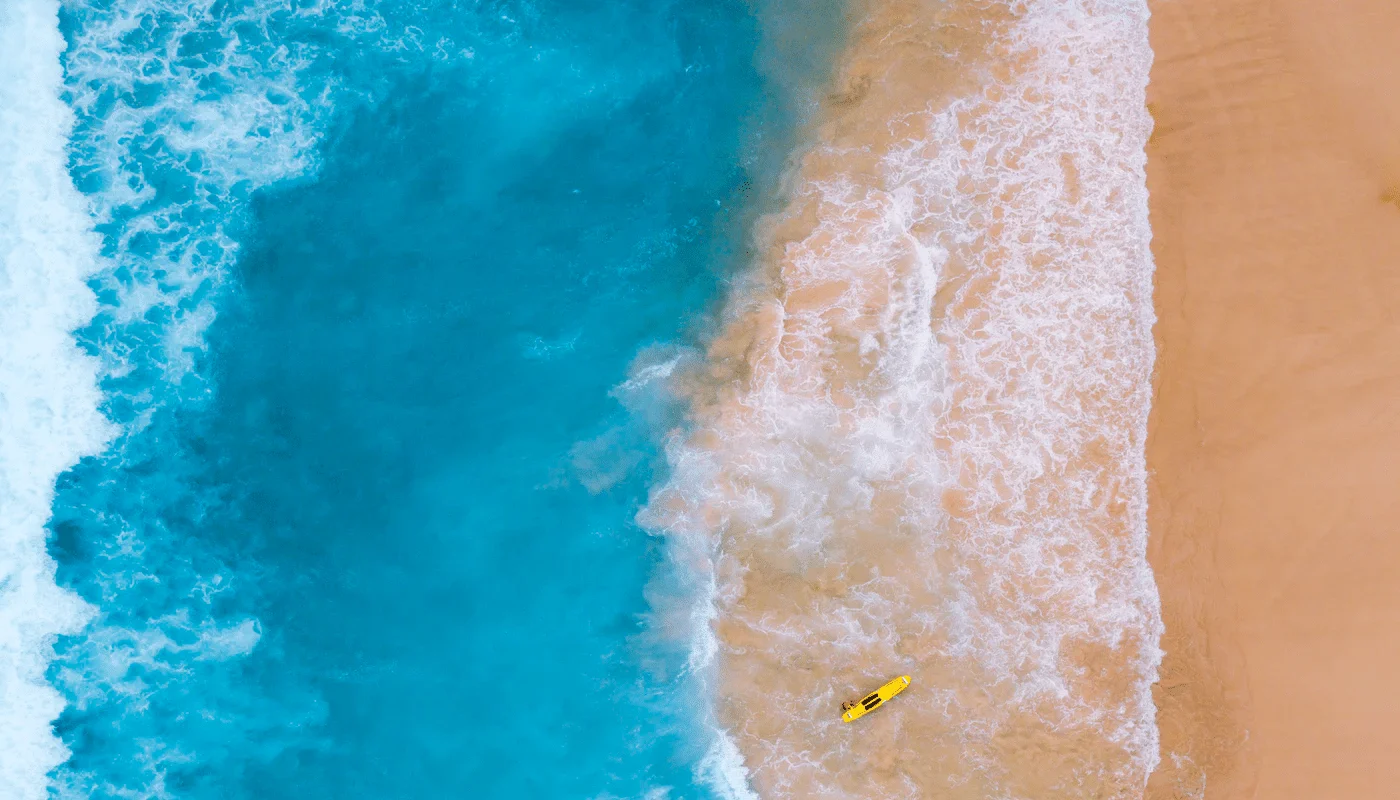 Aerial view of ocean waves meeting sandy beach with yellow surfboard. Beach scene with turquoise water.