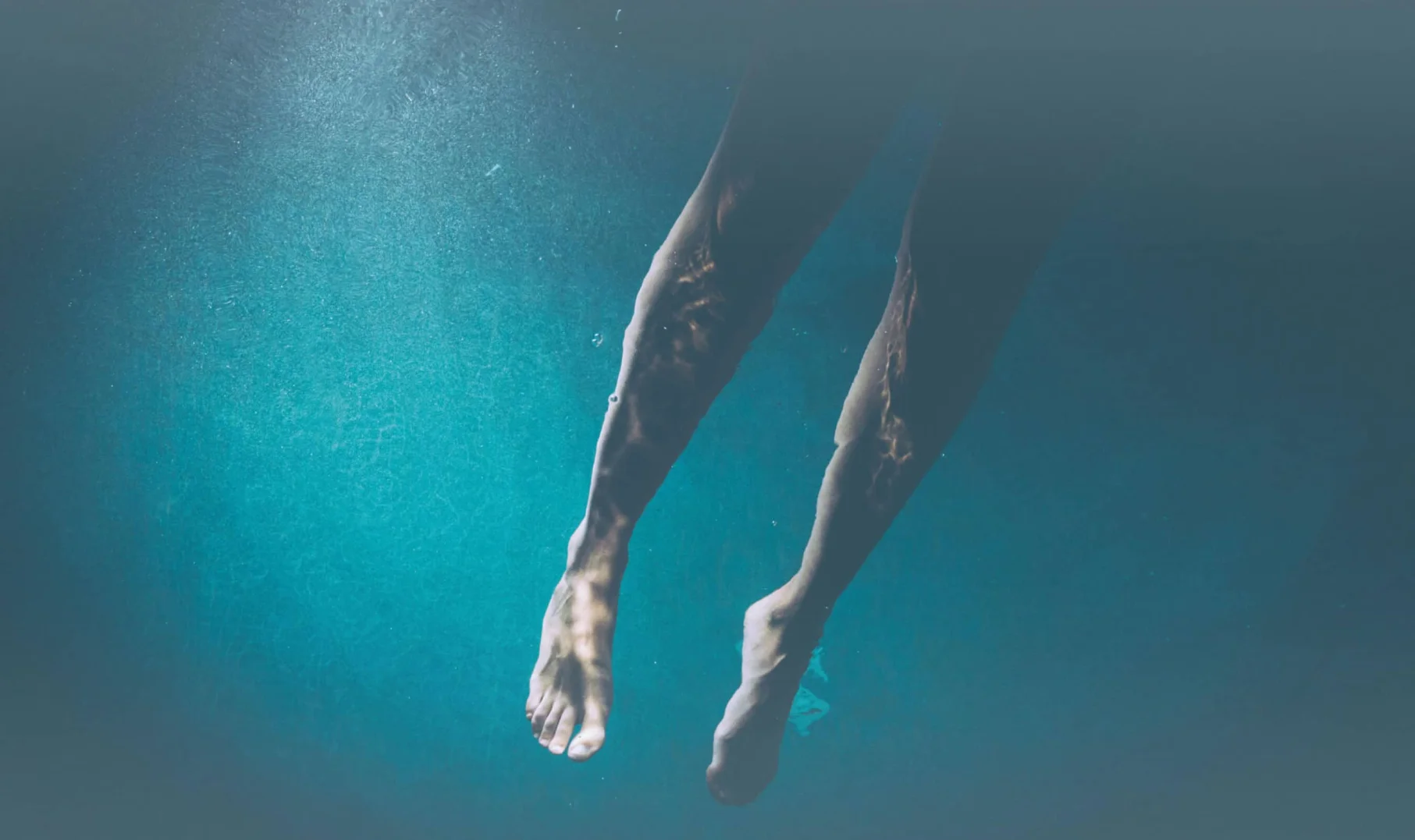 Media: Legs and feet underwater in a blue pool. The light creates a rippling pattern on the skin. Swimming, water, and summer themes.