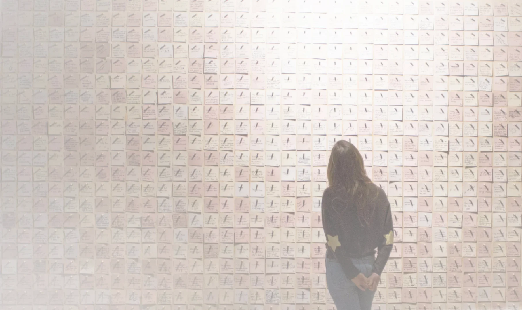 Woman views wall of sticky notes. Art installation with notes. Handwritten messages on display. Interactive art exhibit. Gallery visitor.