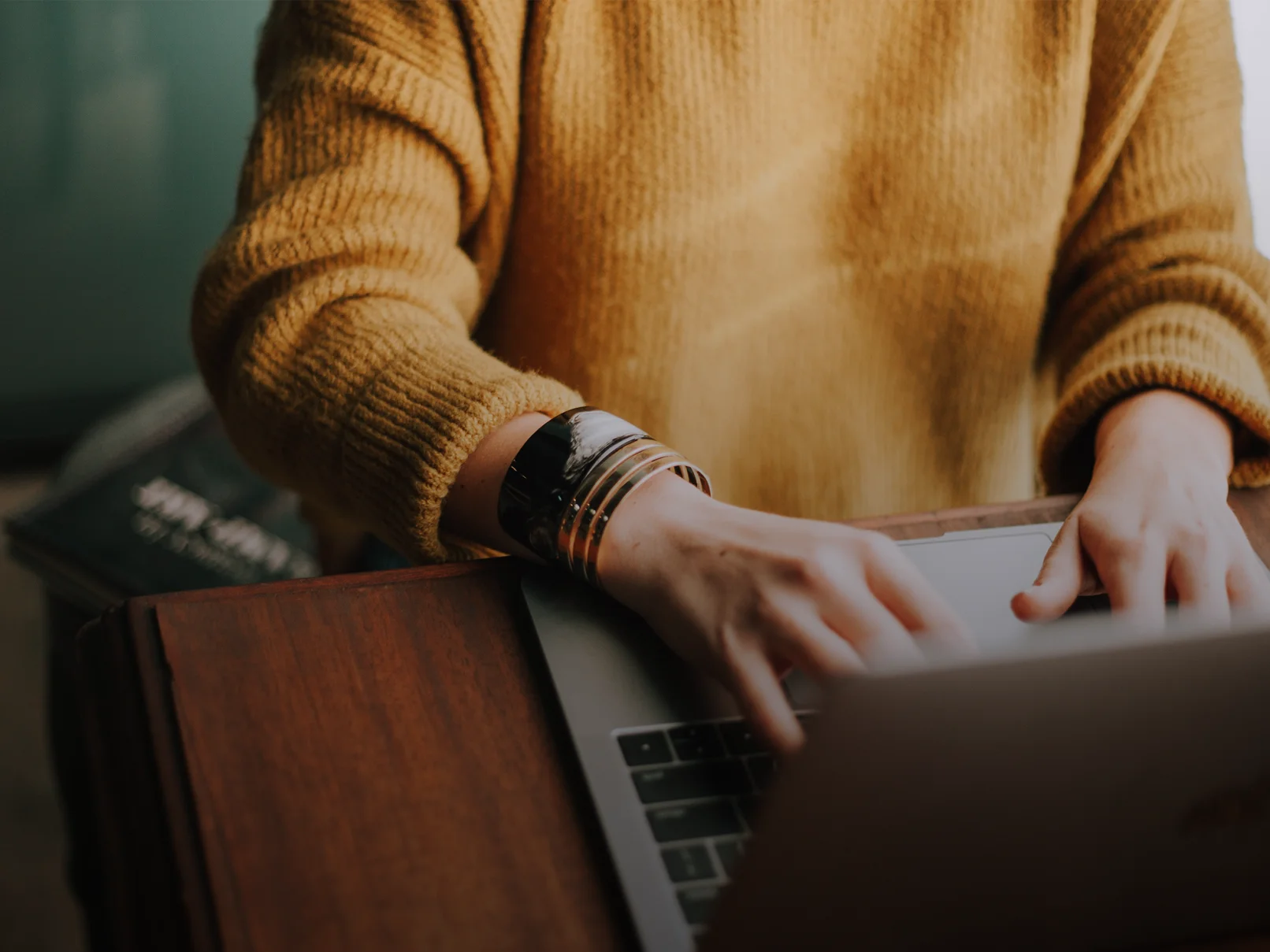 Woman in yellow sweater typing on laptop. Hands with bracelets on keyboard. Wooden desk and blurred background.