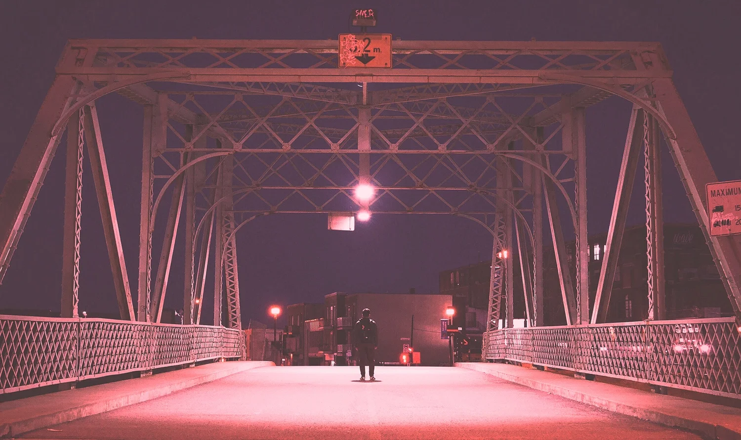 Media: Person stands on pink-lit bridge at night. The steel bridge has a 5.2m clearance sign. City buildings are visible in the background.