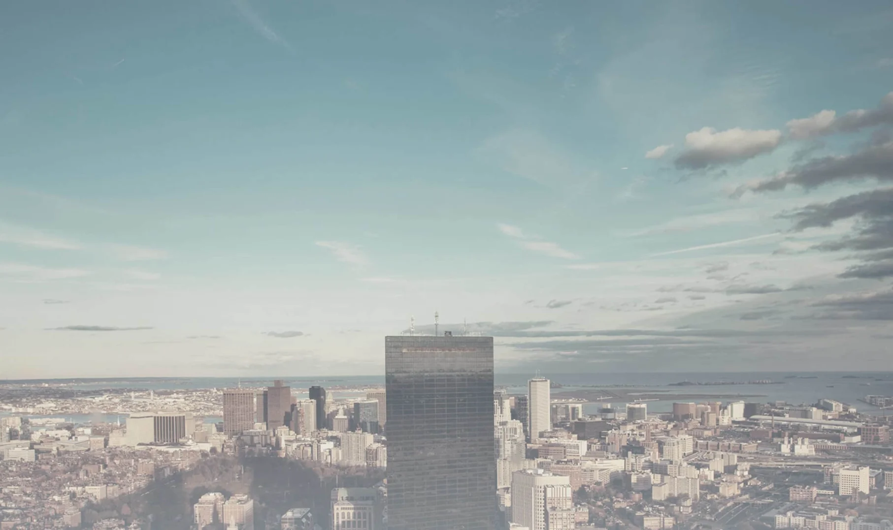 Media: Cityscape view featuring a tall skyscraper, smaller buildings, and a blue sky with scattered clouds. Urban landscape with a muted, vintage aesthetic.