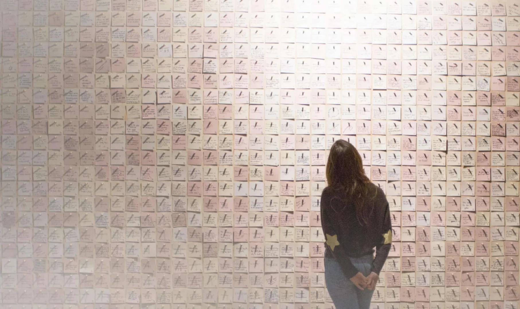 Media: Woman views a wall covered in notes. The notes are pink and have writing on them. The woman is wearing a black shirt and blue jeans.