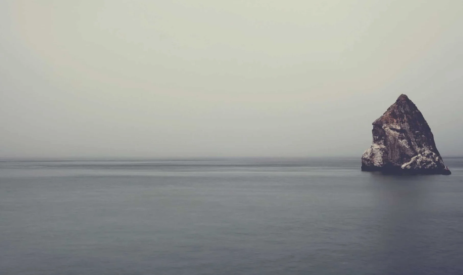 Gloomy seascape featuring a rock formation emerging from the sea under a misty sky. The water is calm, creating a serene yet melancholic scene.