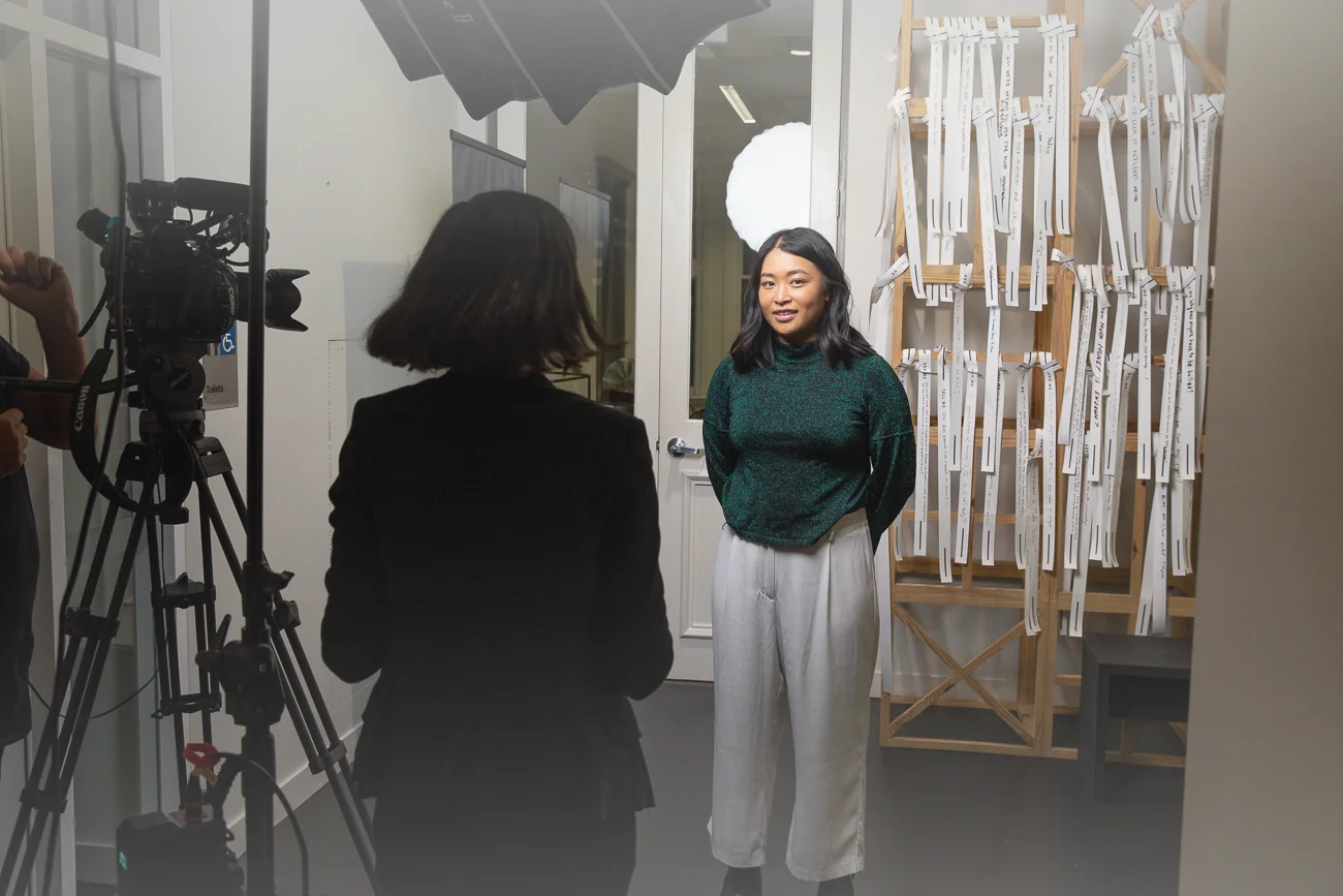 Behind-the-scenes shot of a video interview. A woman is being interviewed by another woman with a camera. Lighting equipment is visible.