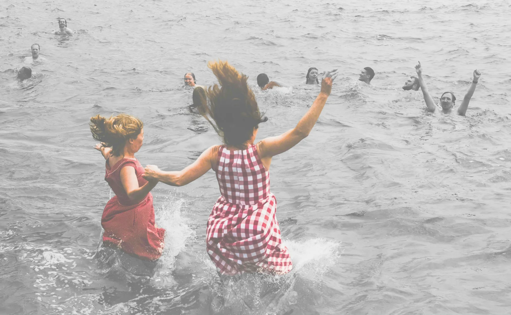 Two women in dresses joyfully run into the water, holding hands. Others swim and play in the background, enjoying a refreshing day at the beach.